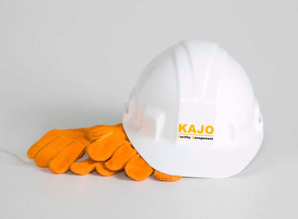 Oranssit työhanskat ja valkoinen kypärä harmaalla taustalla. Kypärässä Kajon logo.